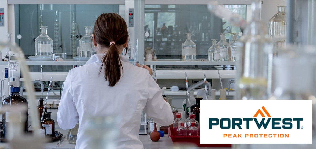 Eine Frau mit dunkelbraunem, zusammengebundenem Haar trägt einen weißen Laborkittel und arbeitet in einem modernen Labor mit verschiedenen Laborgeräten und Chemikalienflaschen. Unten rechts im Bild befindet sich das Logo von "Portwest Peak Protection".