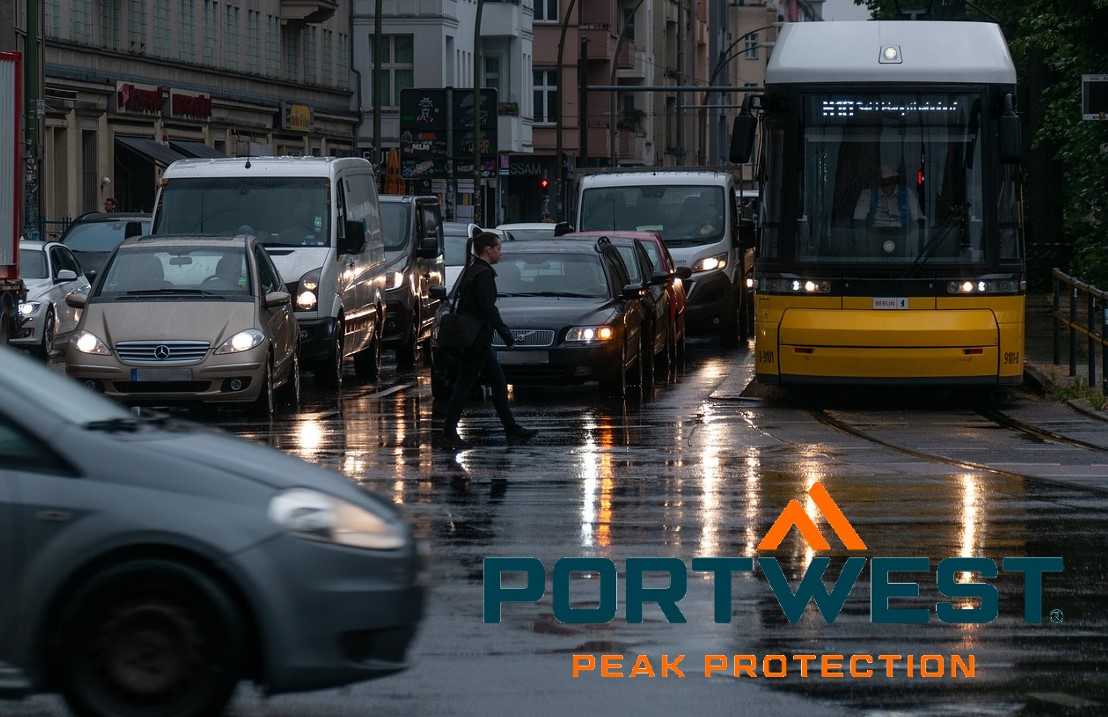 Straßenszene im Regen. Zahlreiche Autos und Busse stehen mit angeschalteten Scheinwerfern an einer Kreuzung. Das Logo der Firma Portwest in Orange und Blau befindet sich im rechten unteren Bildrand. Hinterlegt ist ein Link, der zu unseren Regenjacken führt.