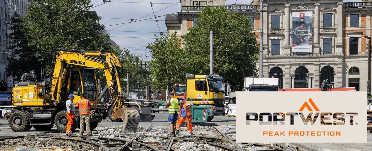 Bauarbeiter in orangefarbener Sicherheitskleidung arbeiten an einer Baustelle in der Stadt. Im Hintergrund sind Bagger, Lastwagen und ein historisches Gebäude zu sehen. Das Portwest-Logo und der Slogan "Peak Protection" sind unten im Bild eingeblendet.