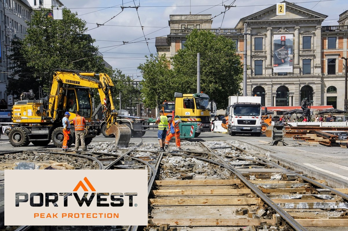 Bauarbeiter in orangefarbener Sicherheitskleidung arbeiten an einer Baustelle in der Stadt. Im Hintergrund sind Bagger, Lastwagen und ein historisches Gebäude zu sehen. Das Portwest-Logo und der Slogan "Peak Protection" sind unten im Bild eingeblendet. Hinterlegt ist ein Link, der zur Artikelseite der Kühlmütze führt.